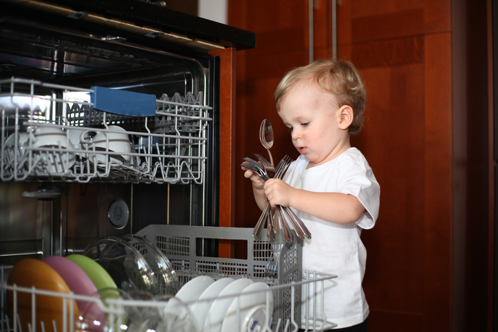 Little boy standing near the dishwasher taking forks and spoons out of it. Playing with cutlery. Baby boy and dishwasher. Baby proofing. Dishwasher with an open door and little boy playing with it. Little helper. Danger of household items for babies.